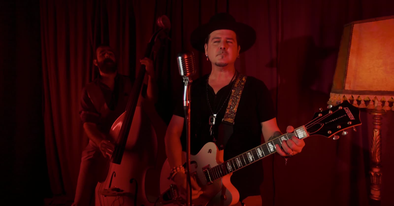 Erik López cantando con una guitarra hollow-body blanca y un contrabajo detrás, bajo luz roja, en el videoclip de Debo partir